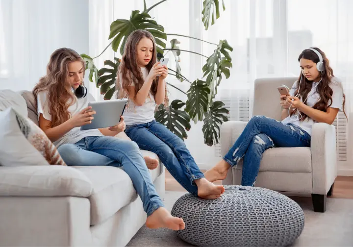 Zdjęcie przedstawia dziewczynki siedzące w salonie i patrzące w ekran urządzeń mobilnych